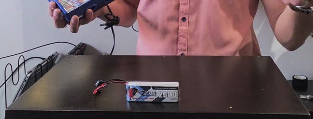 La pila de 2 celdas y 2500 mAh del video sobre la mesa negra, y en mi mano el cargador